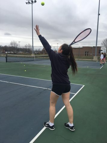 A tennis player has her arms up with a racket in her left hand and is about to hit a serve