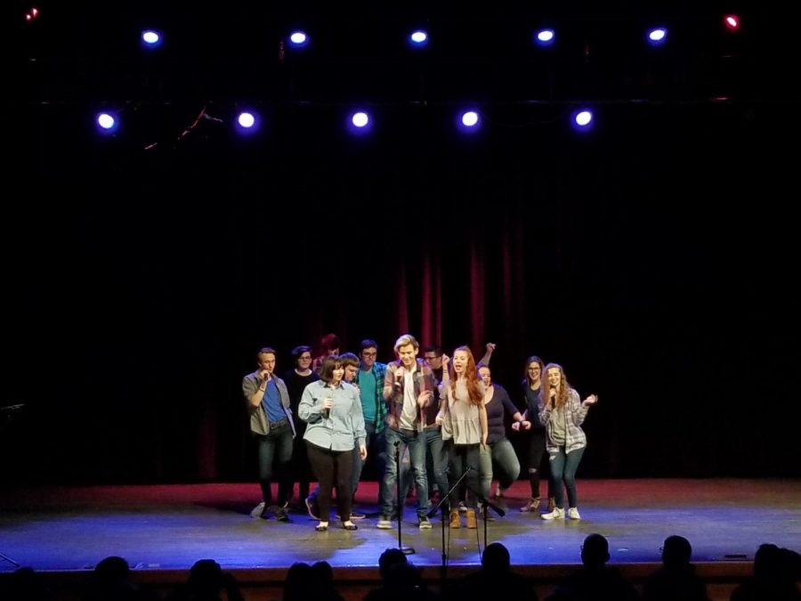 A cluster of performers stand onstage, singing and dancing.