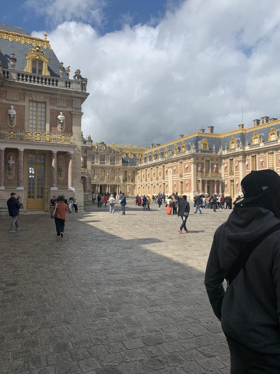 The entrance to Versailles is commonly busy as the weather warms up. The building itself is lined with gold and concrete artistic designs. 
